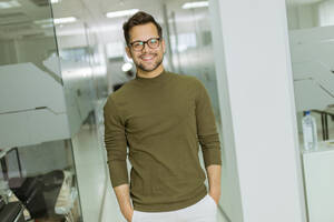 Handsome smiling young businessman in glasses standing in the modern office - INGF13238