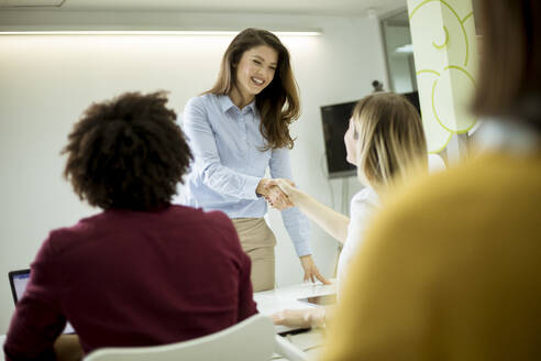 Smiling young woman standing near whiteboard and shaking hand to her female colleague in the small startup office - INGF13250