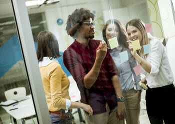 Group of young business people discussing in front of glass wall using post it notes and stickers at startup office - INGF13256