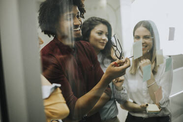 Group of young business people discussing in front of glass wall using post it notes and stickers at startup office - INGF13257