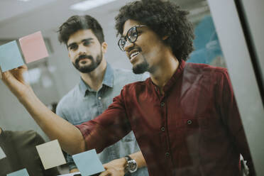 Group of young businessmen discussing in front of glass wall using post it notes and stickers at startup office - INGF13262