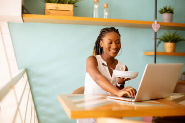 Pretty young black woman having a healthy breakfast while working on laptop in the cafe - INGF13267