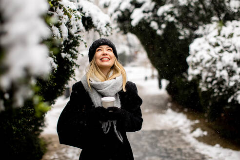 Pretty young woman in warm clothes enjoying in snow with takeaway coffee cup - INGF13270