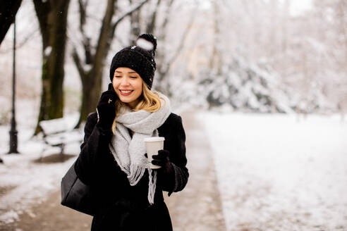A young woman wearing warm winter clothes and a knit hat smiles happily as she stands in the snow and using mobile phone whil holding cofee cup - INGF13272