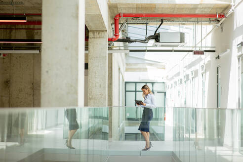 Young business woman with digital tablet walking in the modern office wallway - INGF13291