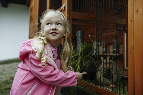 Blondes Mädchen in rosa Regenmantel füttert Kaninchen im Käfig - NSTF00024