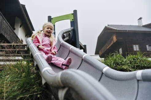 Mädchen im rosa Regenmantel rutscht auf dem Spielplatz die Rutsche hinunter - NSTF00025