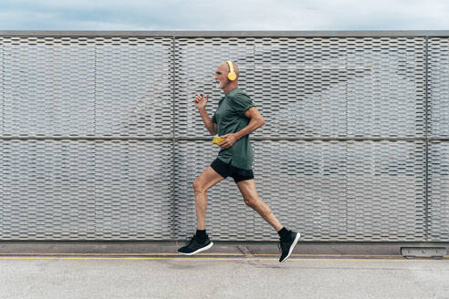 Retired senior man listening to music and running by fence - OIPF04075