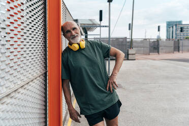 Aktiver älterer Mann mit drahtlosen Kopfhörern am Zaun lehnend - OIPF04077