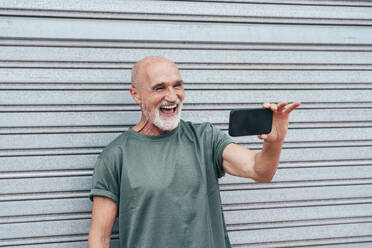 Älterer Mann im Ruhestand lacht bei einem Videogespräch über sein Smartphone vor einem grauen Fensterladen - OIPF04088