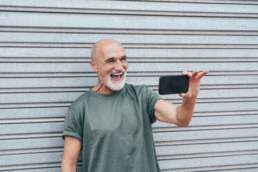 Retired senior man laughing on video call through smart phone in front of gray shutter - OIPF04088