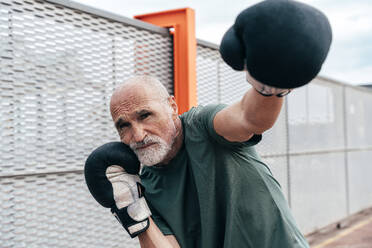 Confident senior man practicing with boxing gloves by fence - OIPF04108