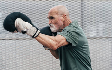 Confident active senior man practicing with boxing gloves in front of fence - OIPF04110