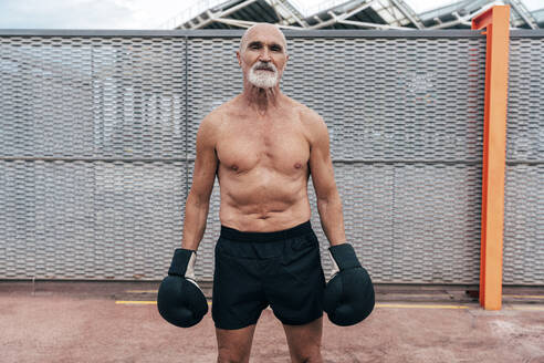 Hemdloser älterer Mann mit Boxhandschuhen vor einem Zaun stehend - OIPF04114