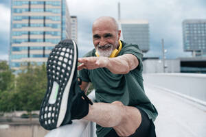 Smiling senior man stretching leg on railing at footbridge in city - OIPF04127