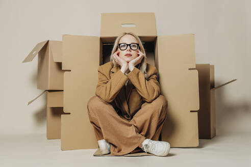 Thoughtful businesswoman sitting near cardboard box against white background - VSNF01715