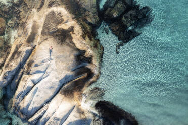 Luftaufnahme eines Mannes in roten Hosen, der bei Sonnenuntergang auf einem Felsen am Meer sitzt, mit kristallklarem tropisch blauem Wasser, Kapstadt, Westkap, Südafrika. - AAEF26495