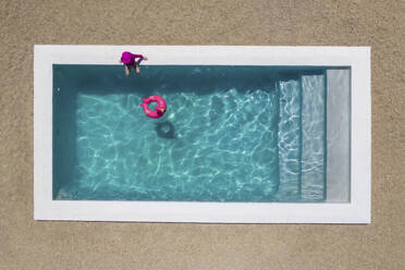 Luftaufnahme eines Mädchens in einem rosafarbenen Kostüm, das an einem Pool mit klarem blauen Wasser und einem rosafarbenen Flamingo-Schwimmkörper im Pool sitzt, Kapstadt, Westkap, Südafrika. - AAEF26509