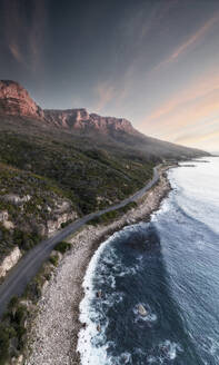 Aerial view of a coastal winding road with mountains on one side and a rocky shoreline on the other, with waves and blue water, Cape Town, Western Cape, South Africa. - AAEF26510