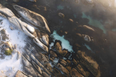 Aerial view of a white sandy beach surrounded by boulders with crystal clear blue water and fog creating a warm haze at sunset, Cape Town, Western Cape, South Africa. - AAEF26521