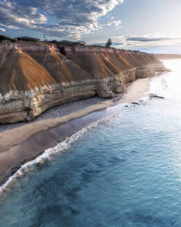 Aerial view of a man in an orange jacket walking on a beach with limestone cliffs towering over him and blue ocean water lapping the shoreline, Port Willunga, South Australia, Australia. - AAEF26541