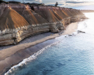 Luftaufnahme eines Mannes in einer orangefarbenen Jacke, der an einem Strand spazieren geht, an dem sich Kalksteinklippen erheben und blaues Ozeanwasser an die Küste plätschert, Port Willunga, South Australia, Australien. - AAEF26542