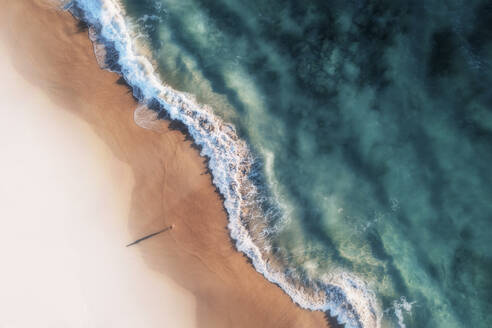 Luftaufnahme eines weißen Sandstrandes mit weißen Wellen, die ans Ufer rollen, und kristallklarem, blauem Wasser und einem Mädchen, das am Strand steht und einen langen Schatten wirft, Port Noarlunga, South Australia, Australien. - AAEF26544