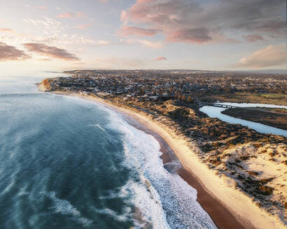 Luftaufnahme eines langen Sandstrandes bei Sonnenuntergang, mit blauen Wellen, die an das Ufer rollen, einem Fluss und rosafarbenen Abendwolken im Hintergrund, Port Noarlunga, South Australia, Australien. - AAEF26553