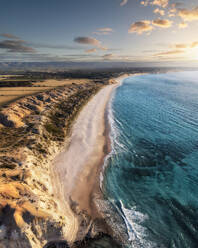 Aerial view of a white sandy beach edged with limestone cliffs and blue water at sunset with some clouds in the sky and farm fields at the top of the cliffs, Port Willunga, South Australia, Australia. - AAEF26558