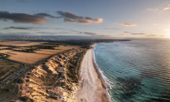 Luftaufnahme eines weißen Sandstrandes mit Kalksteinklippen und blauem Wasser bei Sonnenuntergang mit einigen Wolken am Himmel und landwirtschaftlichen Feldern an der Spitze der Klippen, Port Willunga, South Australia, Australien. - AAEF26559