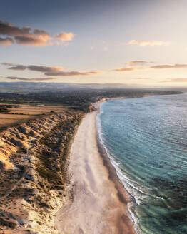 Aerial view of a white sandy beach edged with limestone cliffs and blue water at sunset with some clouds in the sky and farm fields at the top of the cliffs, Port Willunga, South Australia, Australia. - AAEF26560