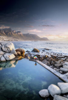 Aerial view of a person standing by a rock pool surrounded by boulders, holding a colorful umbrella and overlooking the ocean waves and some mountains, Cape Town, Western Cape, South Africa. - AAEF26564