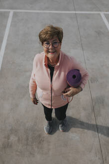 Smiling senior woman standing with exercise mat at parking lot - DMGF01253