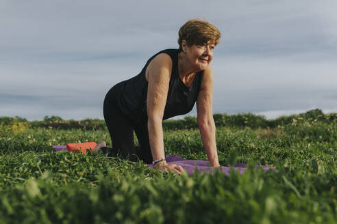 Ältere Frau übt Yoga auf einer Übungsmatte inmitten von Pflanzen auf einer Wiese - DMGF01277