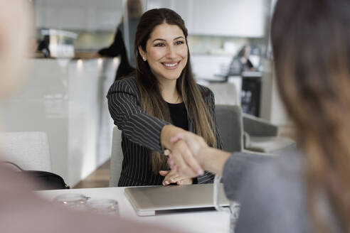 Happy saleswoman greeting female customer with handshake while sitting at desk in real estate office - MASF43554