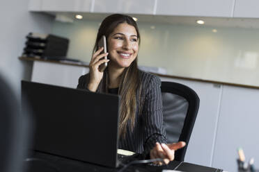 Smiling real estate agent talking on smart phone while working in office - MASF43561