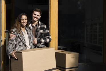 Portrait of smiling man embracing woman holding cardboard box in front of apartment - MASF43581