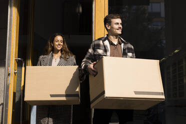 Smiling young couple carrying boxes while walking outside apartment building - MASF43587