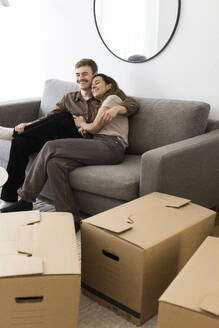 Happy couple embracing while sitting on sofa by cardboard boxes at home - MASF43626