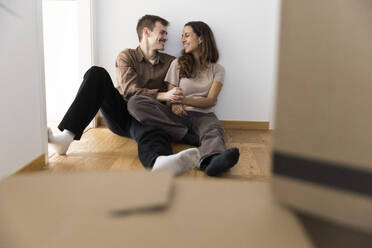 Happy multiracial couple with hands intertwined sitting on hardwood floor at home - MASF43628