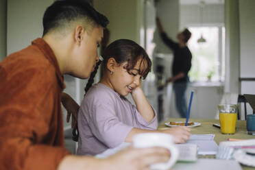 Smiling girl doing homework while sitting with gay father at table in home - MASF43639