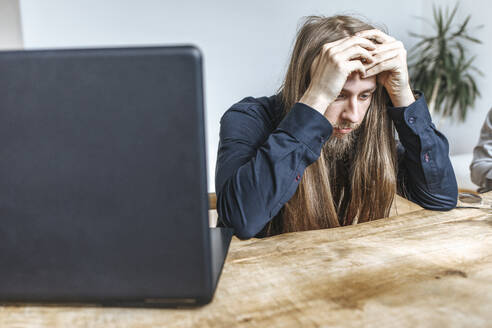 Gestresster Hipster-Programmierer sitzt mit dem Kopf in den Händen am Laptop auf dem Tisch im Büro - EVKF00115