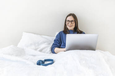 Lächelnder Programmierer mit Laptop, der zu Hause auf dem Bett sitzt - EVKF00120