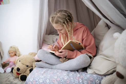 Mädchen mit Zöpfen schreibt in Buch in grauem Baldachin zu Hause - NJAF00824