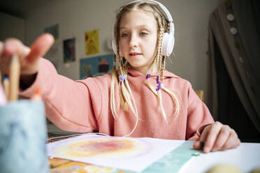 Girl wearing headphones taking pencil from desk organizer at table - NJAF00830