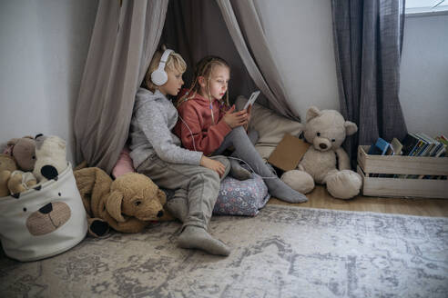 Curious boy with headphones looking at sister using smart phone in gray tent at home - NJAF00834