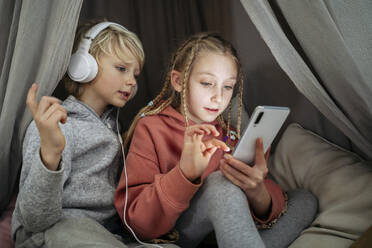 Neugieriger Junge mit Kopfhörern, der seine Schwester bei der Benutzung eines Mobiltelefons im Zelt zu Hause beobachtet - NJAF00836