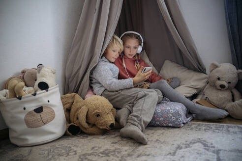 Sister with headphones looking at boy holding smart phone in gray tent at home - NJAF00837