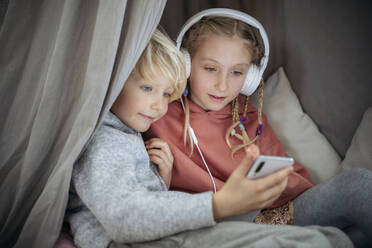 Curious brother and sister looking at smart phone in gray tent at home - NJAF00838