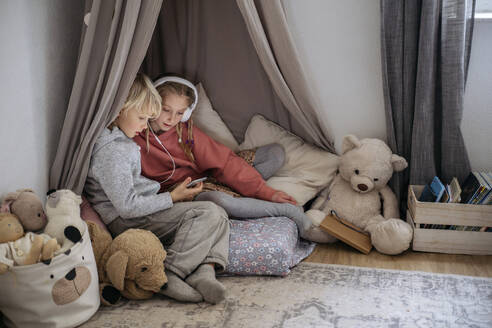 Girl with headphones looking at brother using smart phone in gray tent at home - NJAF00839